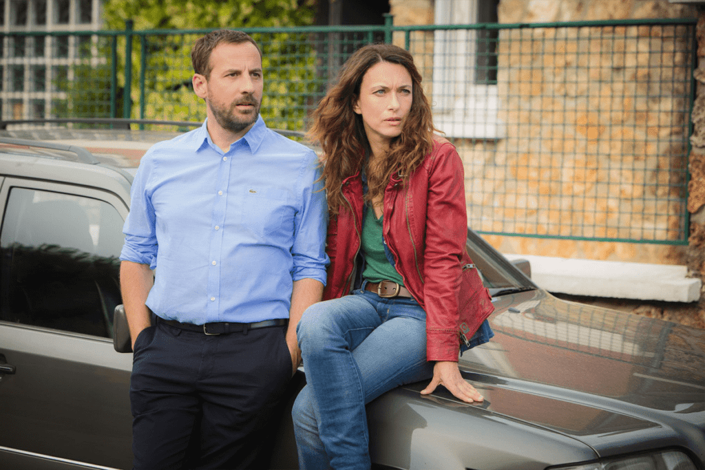 "Natacha Lindinger, connue pour ses scènes nues, captivée par une vision lointaine dans une scène de film, vêtue d'une veste rouge à côté d'un homme, dans une voiture